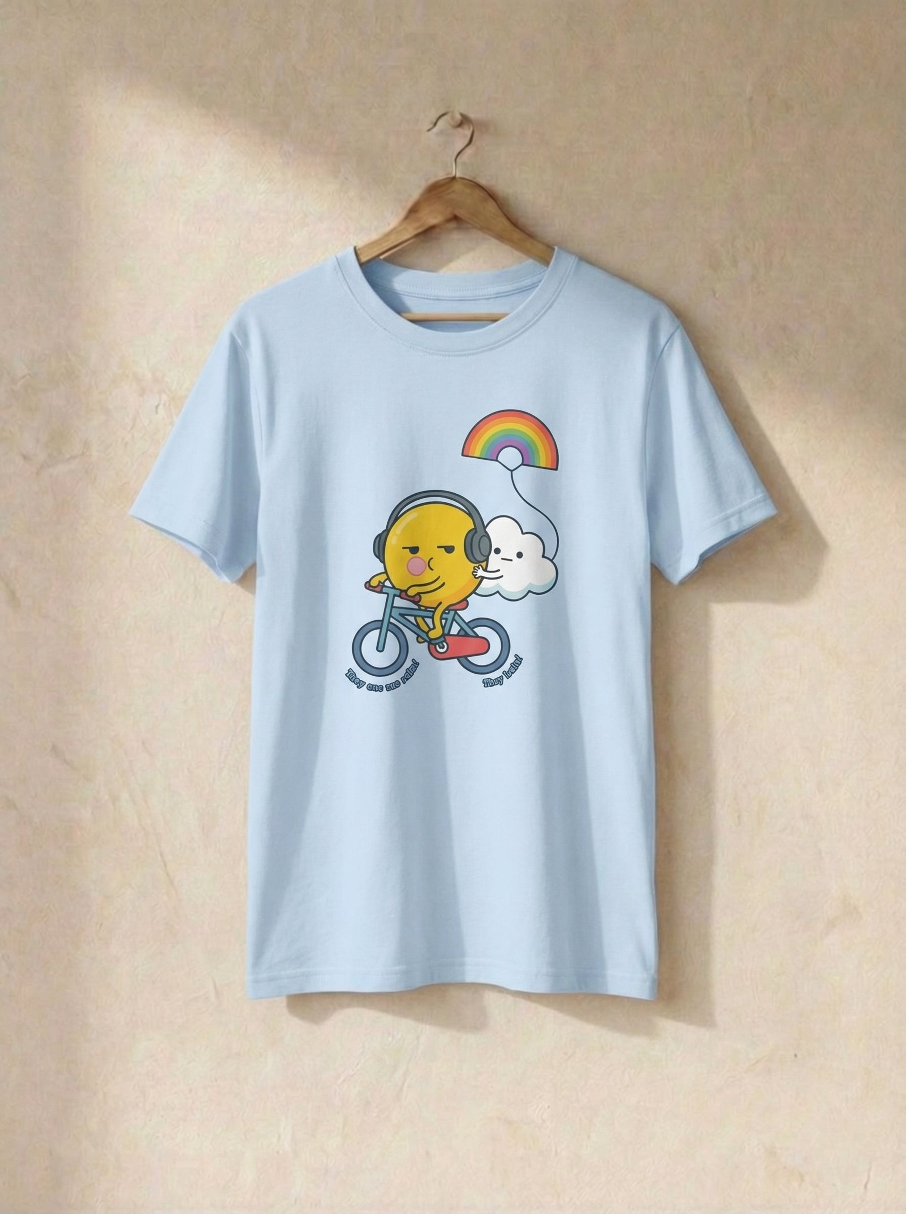 Light blue t-shirt with a cartoon design of a face riding a bike with a rainbow and cloud, on a beige background.