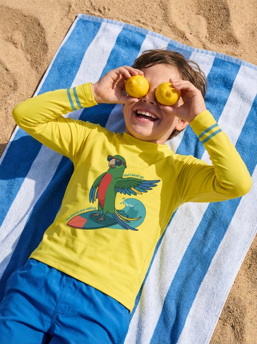 Child wearing a yellow shirt with a parrot design, lying on a blue and white striped towel at the beach.