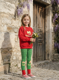 Young girl in red sweater and green patterned pants holding flowers in front of a stone building with wisteria.