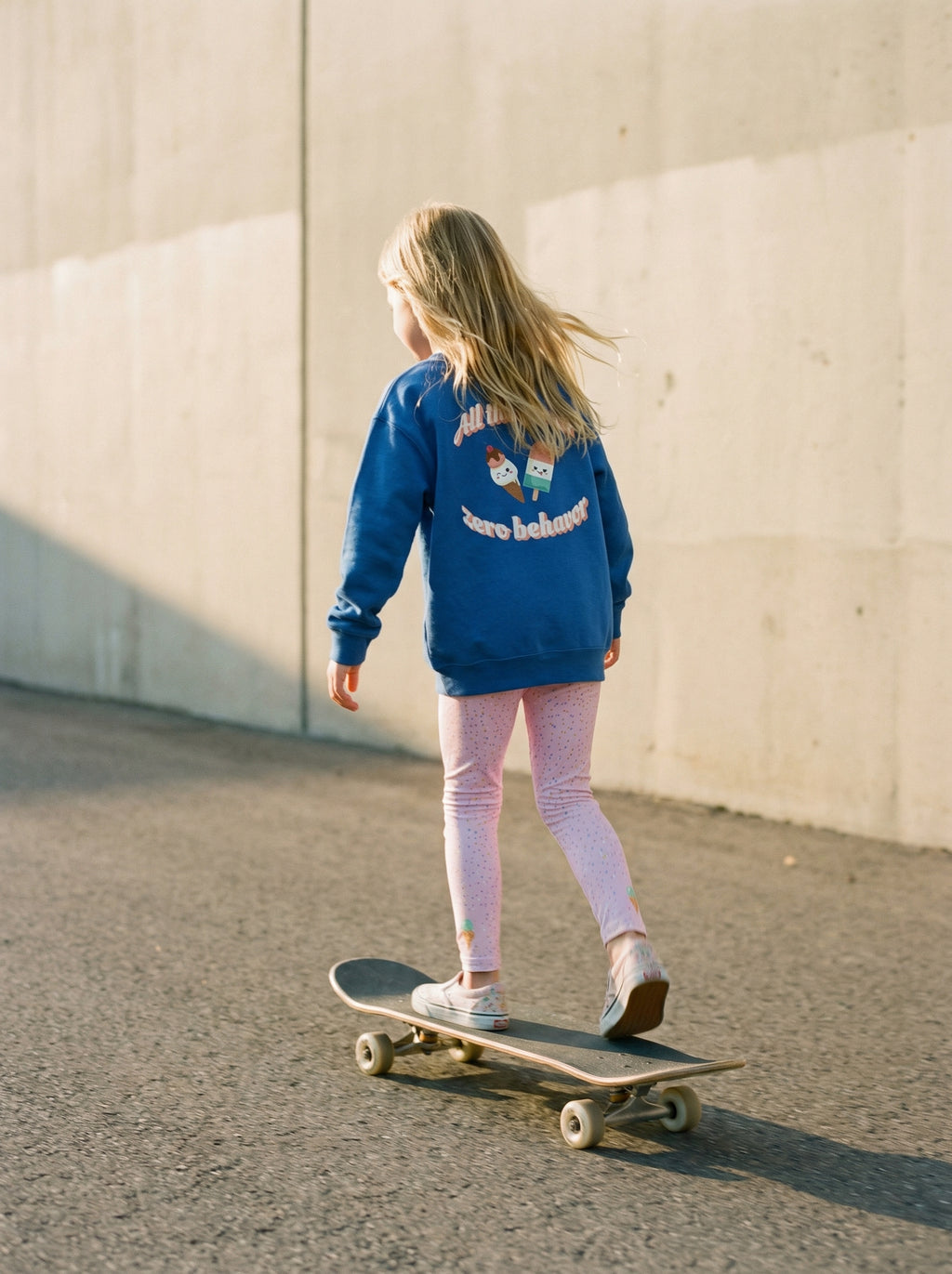 Child in a blue sweatshirt and pink pants standing on a skateboard against a light-colored wall.