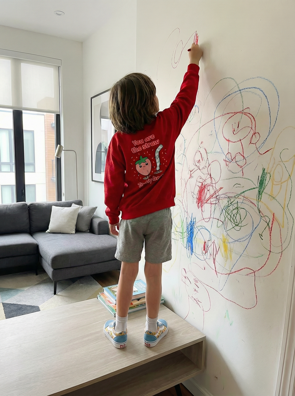 Child in a red sweatshirt drawing on a wall with colorful markers in a living room.