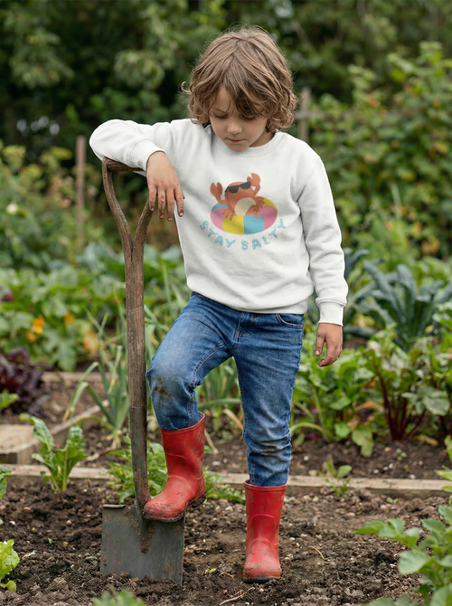Child in a garden wearing a white sweatshirt with a colorful design, blue jeans, and red boots holding a shovel.