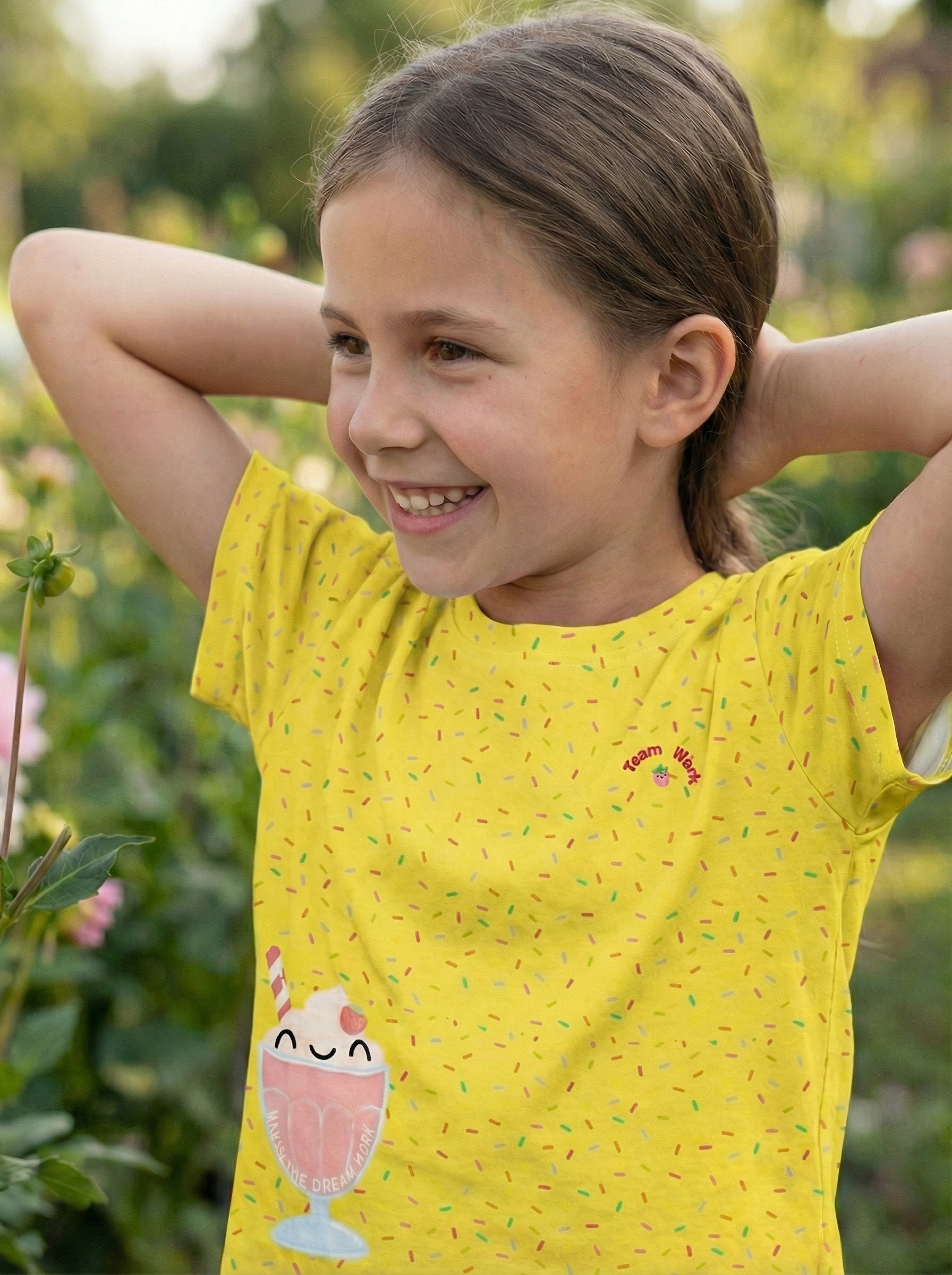 Young girl wearing a yellow shirt with a dessert design, standing outdoors.